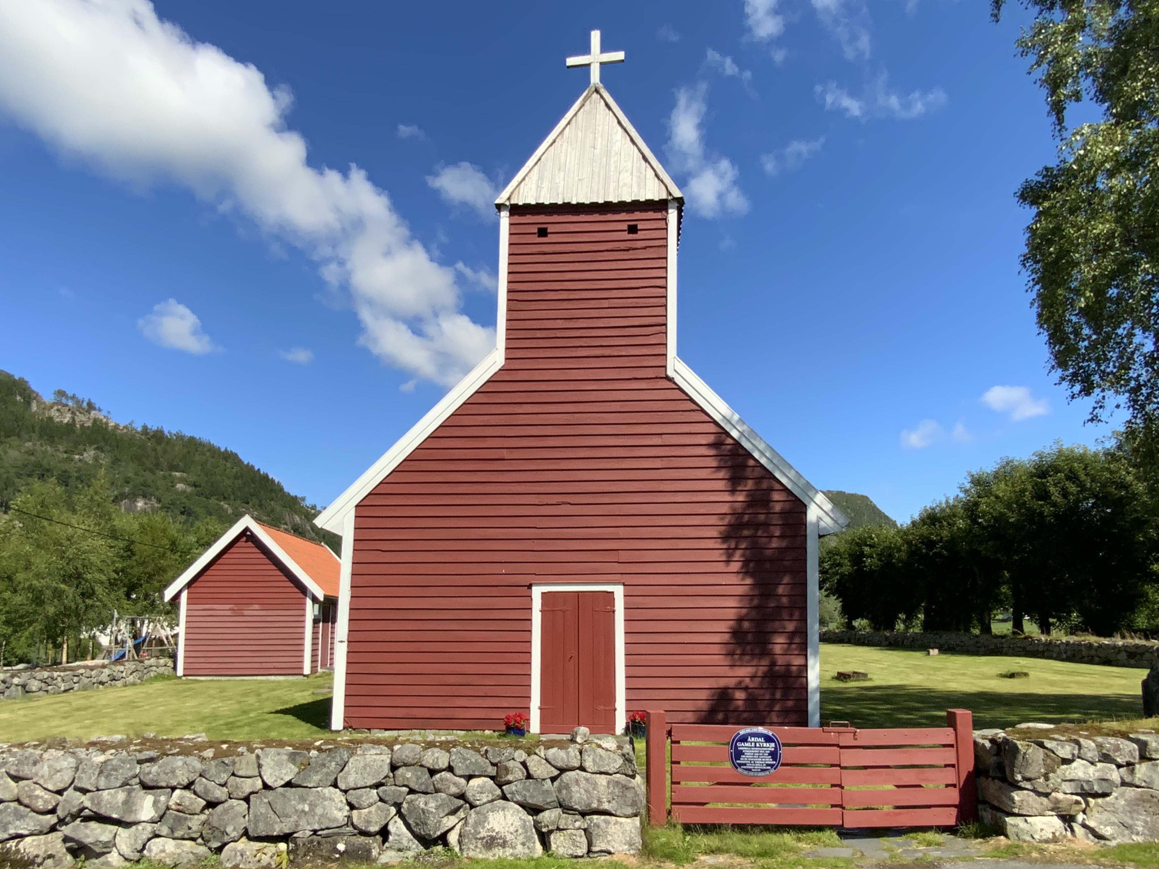Årdal gamle kyrkje i solskinn. Raud trekyrkje med kvitt kors. 